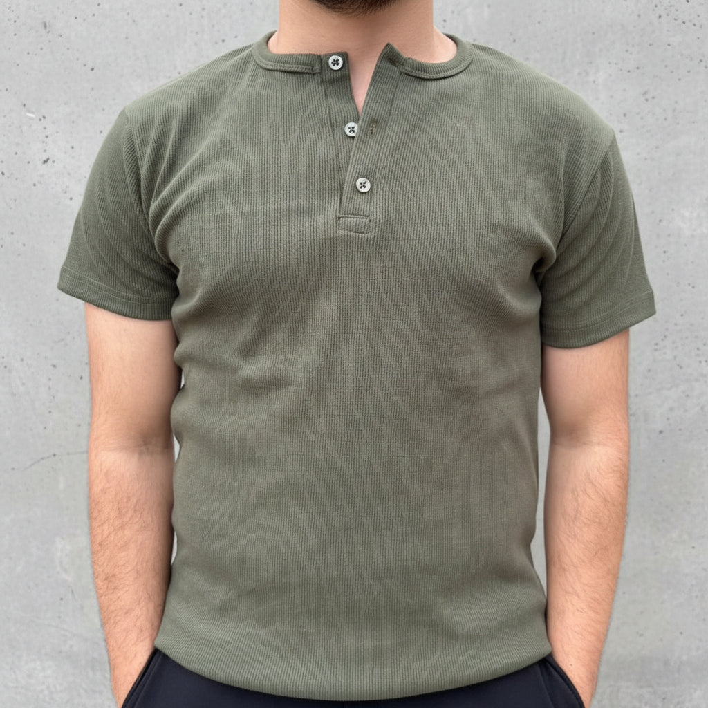 Person wearing a green henley shirt standing outdoors with plants and people in the background